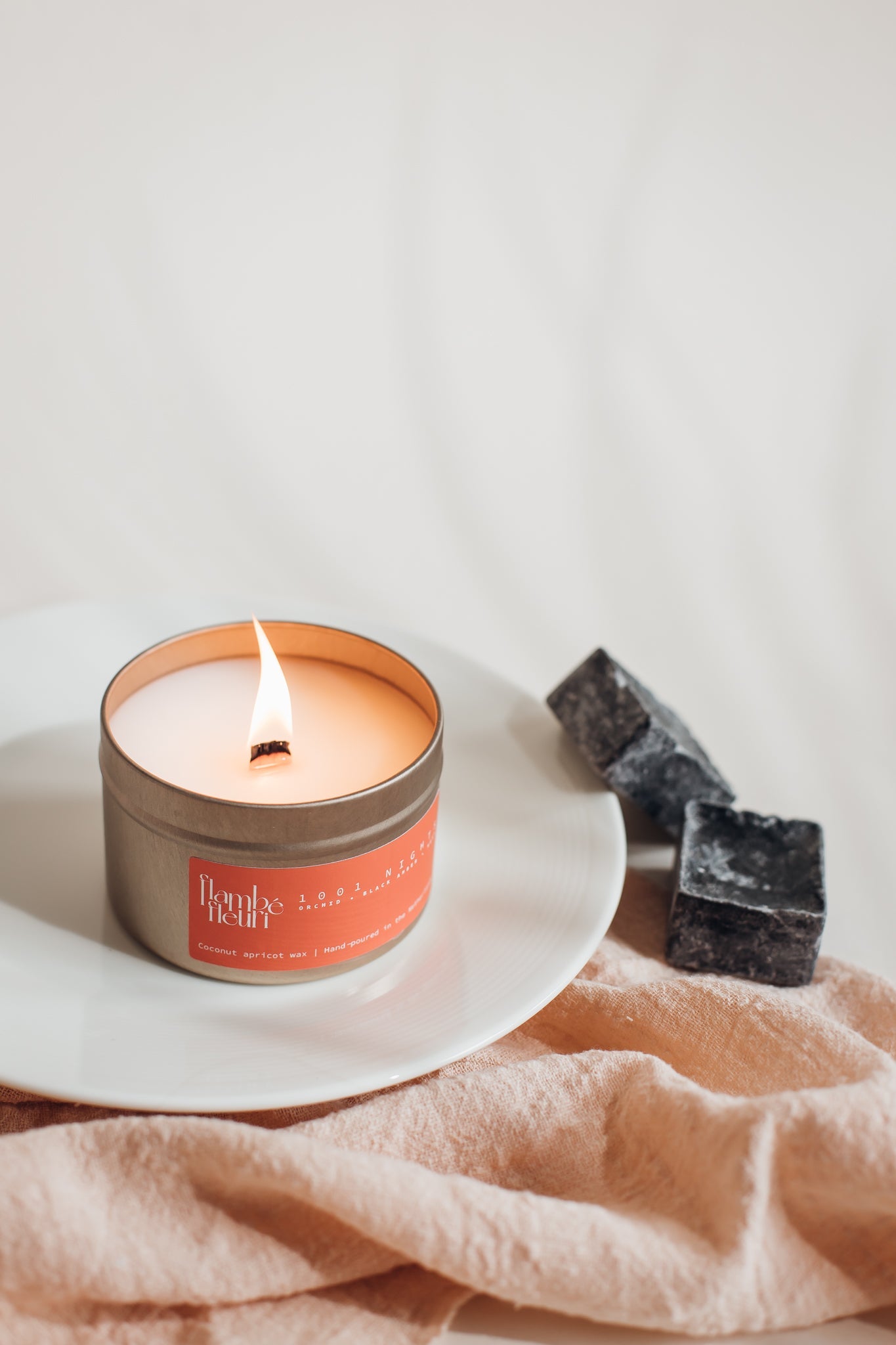 Vegan Scented Candle - Oriental 1001 Nights - Non Toxic. Candle in a tin with a label on a white plate, surrounded by dark soap bars on a light fabric background.