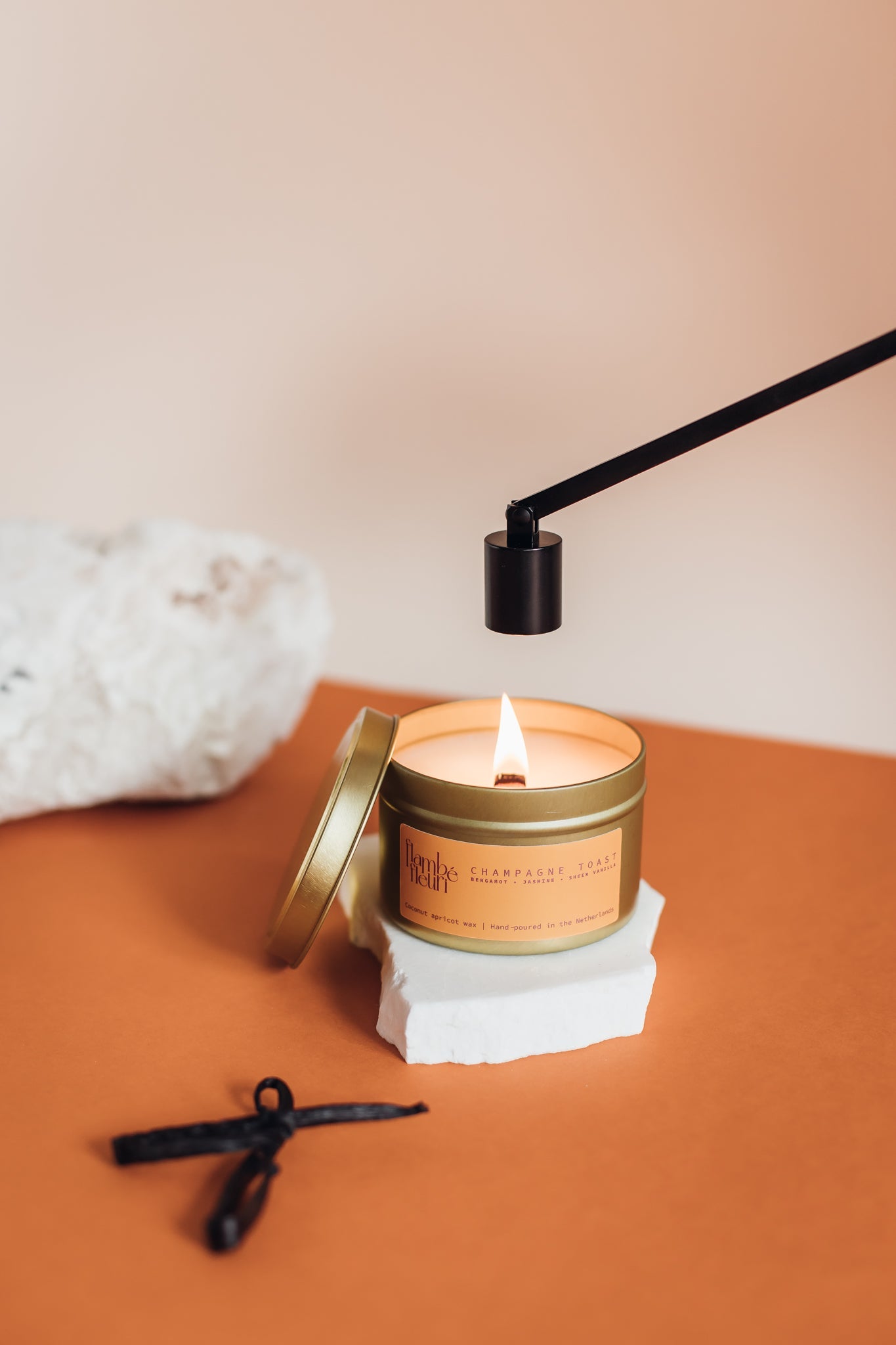 Gold vegan non toxic candle in a tin with a lit wooden wick on a white block, surrounded by decorative items on a peach background.