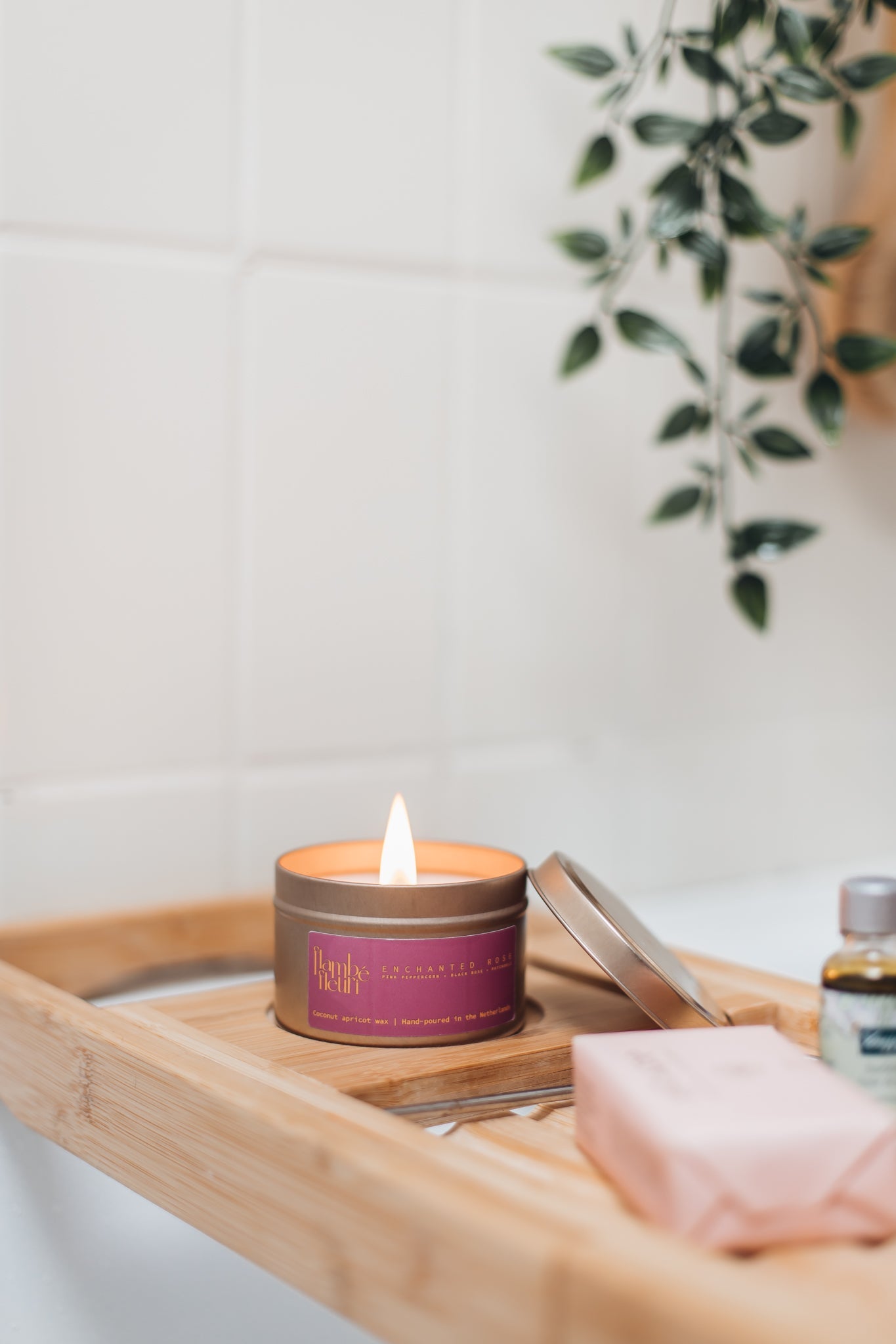 Non toxic vegan Candle in a tin with a purple label on a wooden tray over a bathtub, with a plant in the background. Wooden wick 