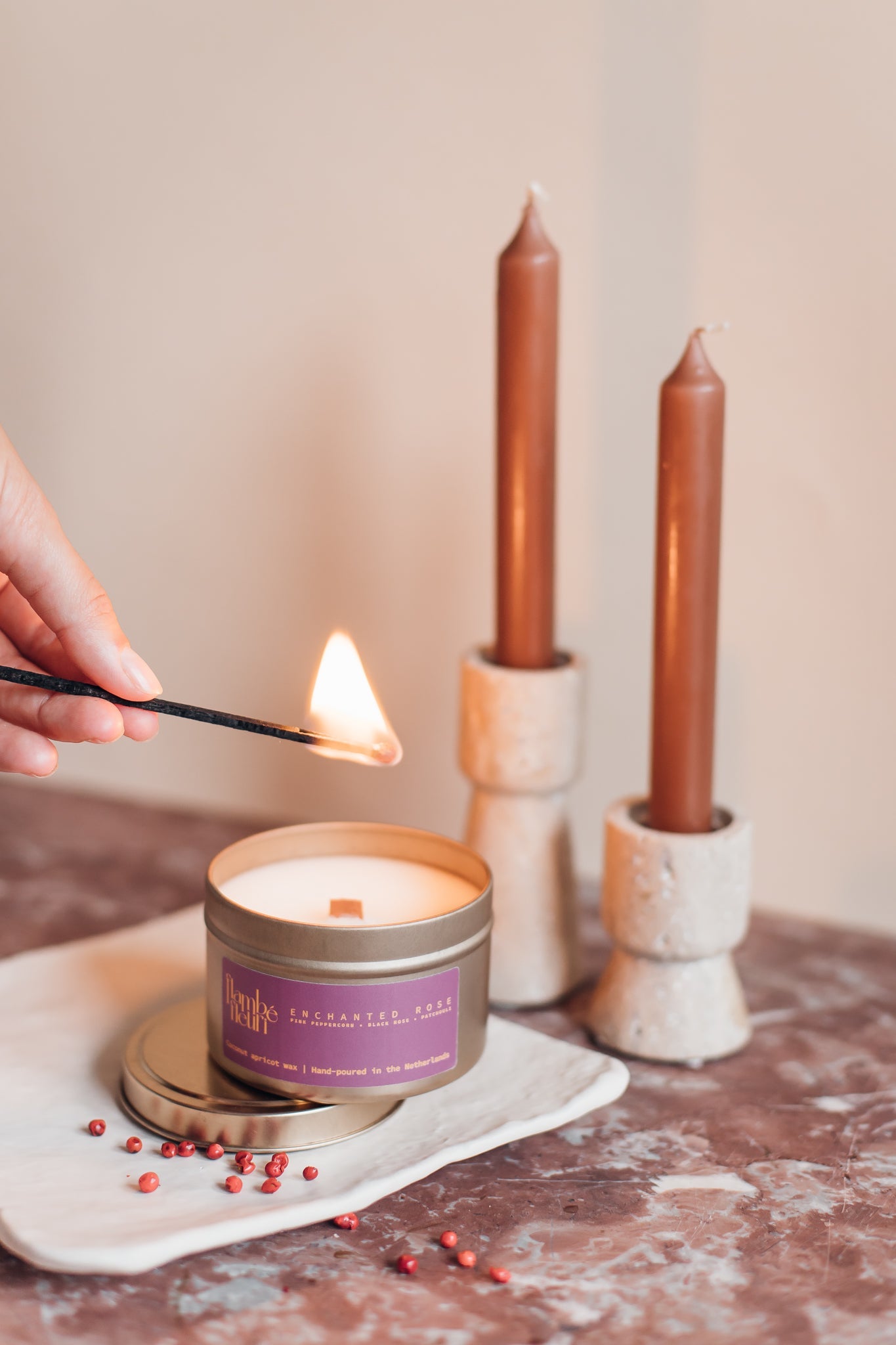 Person lighting a non toxic vegan candle with a match, surrounded by two tall candles in stone holders on a neutral background. Wooden wick 